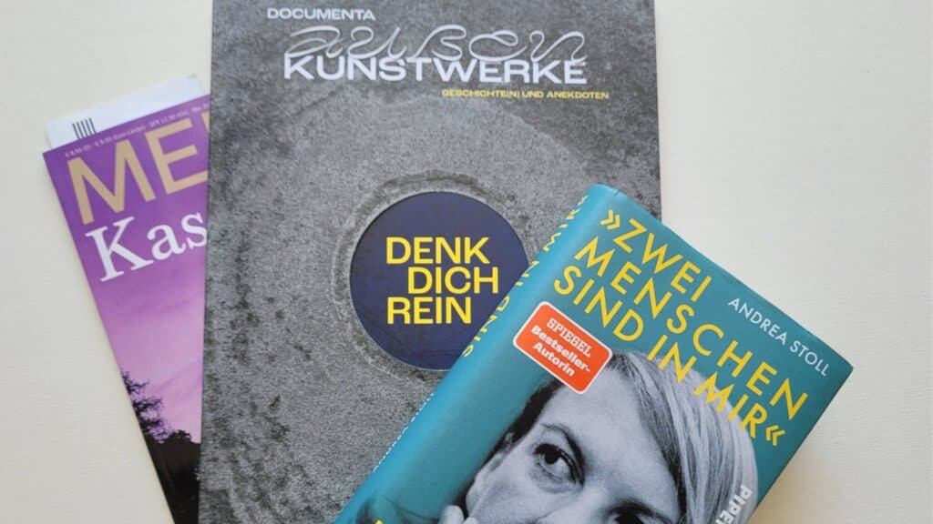 Drei übereinandergelegte Publikationen: eine Zeitschrift, ein Documenta-Booklet und das Buch 'Zwei Menschen sind in mir' von Andrea Stoll.