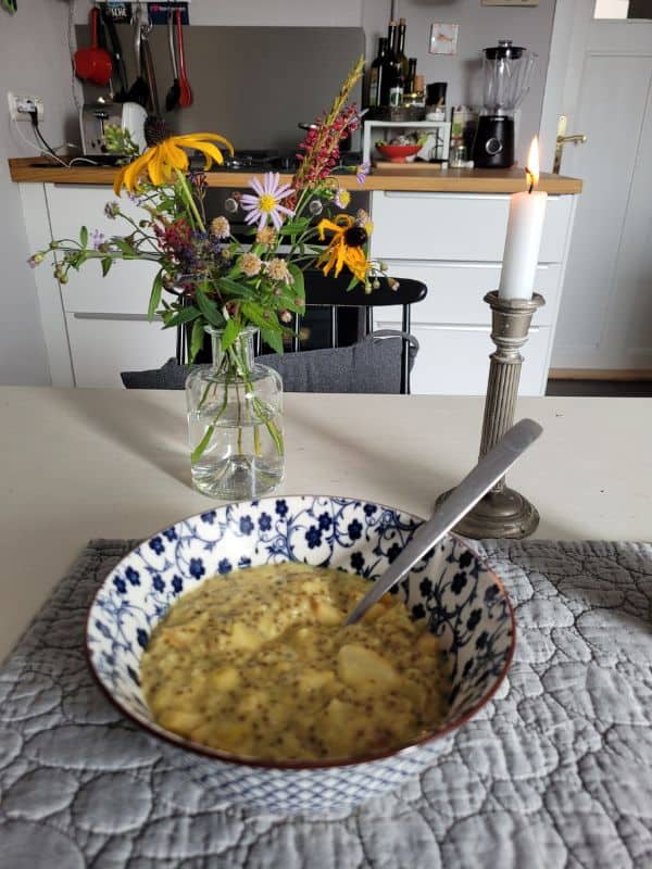 Schale mit Porridge und Löffel auf einem gedeckten Frühstückstisch, im Hintergrund ein bunter Blumenstrauß und eine brennende Kerze