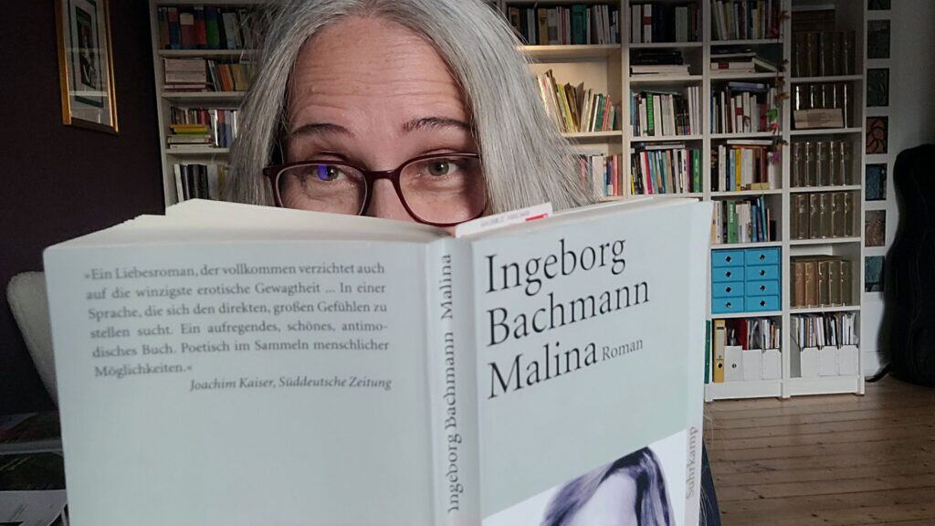 Frau mit roter Brille blickt hinter einem Buch hervor. Im Hintergrund ein prall gefülltes Bücherregal.